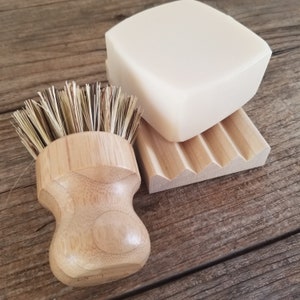 May include: A natural soap bar, wooden soap dish, and a bamboo dish brush are arranged on a rustic wooden surface. The soap is a light cream color, and the dish has a grooved design. The brush has a bamboo handle and natural bristles.