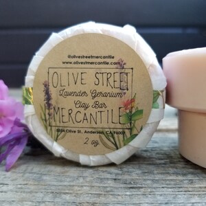 May include: A round, white clay bar wrapped in paper with a brown label that reads "OLIVE STREET Lavender Geranium Clay Bar MERCANTILE 5554 Olive St., Anderson, CA 96007 2 oz."