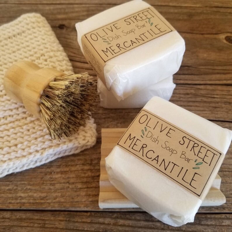 May include: White dish soap bars from Olive Street Mercantile, wrapped in paper, with a brown label. A wooden dish brush and a knitted dishcloth are also present. The soap bars are stacked on a wooden surface.
