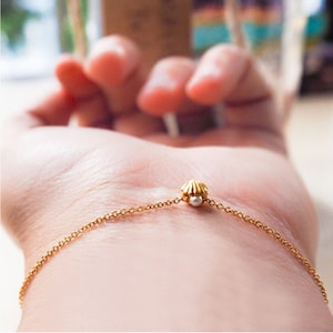 Puede incluir: Un delicado brazalete de cadena dorada con un pequeño dije de concha y un acento de perla.