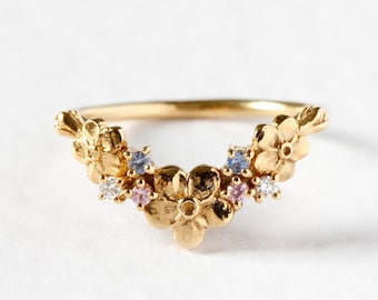 May include: A gold-colored ring featuring a floral design. The ring is adorned with small, colorful gemstones, including pink, blue, and white stones. The ring's band is thin, and the floral arrangement curves across the top.