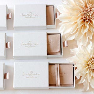 May include: White textured boxes with pull-out drawers, each featuring a gold logo and website address. Inside, a blush-colored pouch is visible. Soft pink ribbon pulls and large cream-colored flowers complete the elegant presentation.