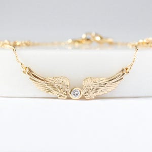 May include: A gold-colored necklace featuring a pair of detailed wings with a small, round, clear gemstone in the center. The wings are slightly curved, and the necklace chain is delicate.
