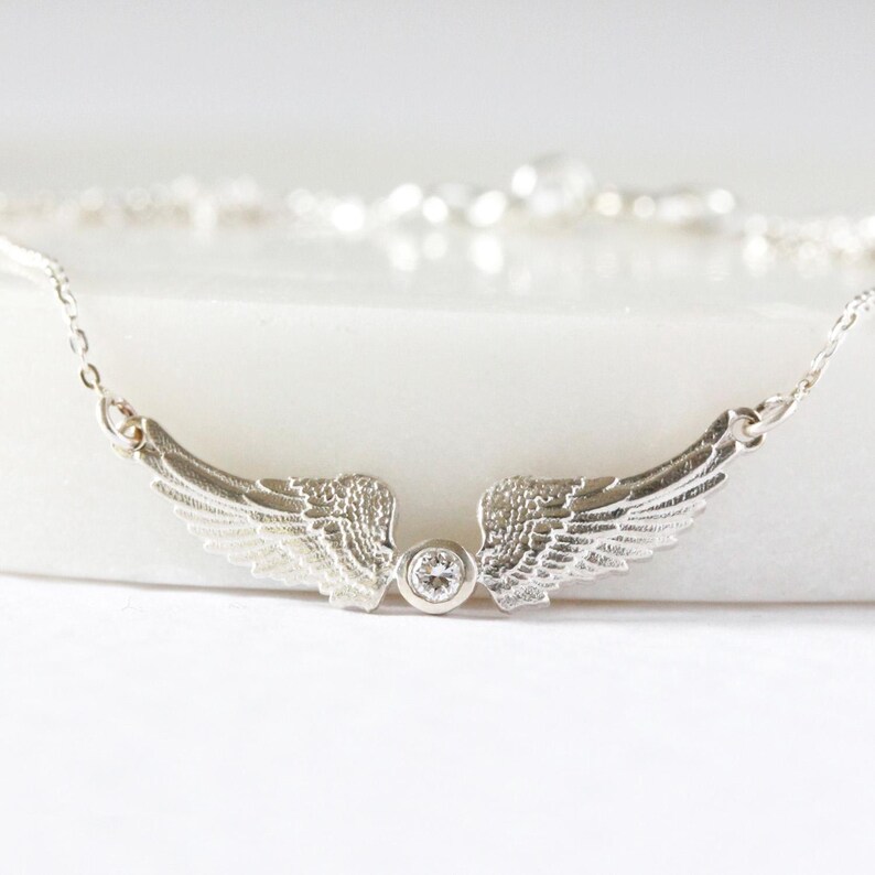 May include: A silver-toned necklace featuring a pair of detailed wings with a central, round, clear gemstone. The wings are intricately designed, and the necklace chain is delicate. The jewelry is set against a white background.