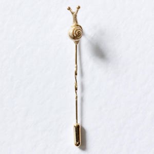 Pode incluir: Um alfinete de ouro em forma de caracol com uma concha detalhada e antenas enroladas. O alfinete é preso a um caule longo, fino e dourado com um design retorcido.
