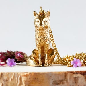 Puede incluir: Un collar con un colgante de zorro dorado. El zorro está sentado con la cola enroscada alrededor de sus patas. El collar está hecho de una cadena dorada.