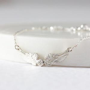 May include: A silver-colored bracelet featuring a pair of detailed, textured wings. A small, round, clear gemstone is set between the wings. The bracelet is on a white surface.
