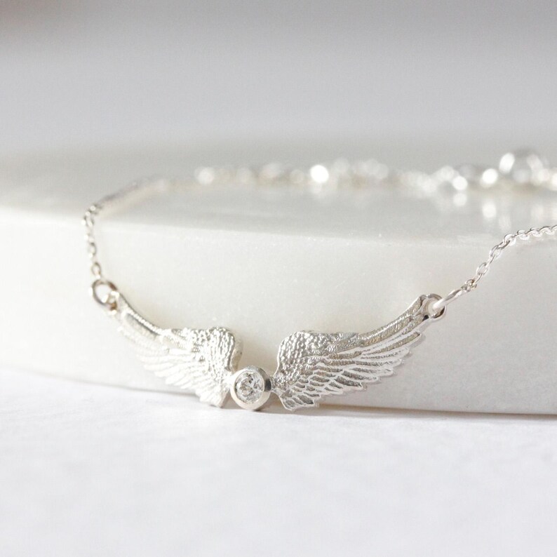 May include: A silver-colored bracelet featuring a pair of detailed, textured wings. A small, round, clear gemstone is set between the wings. The bracelet is on a white surface.