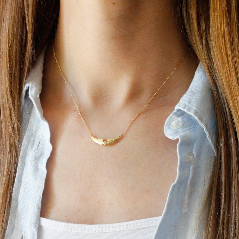 May include: A delicate gold-colored necklace featuring a wing design with a small, clear gemstone in the center. The necklace is worn against a light blue shirt and white top, creating a subtle contrast.