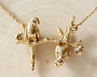 May include: Gold necklace featuring two parrots perched on a branch. The parrots are detailed and realistic, with textured feathers. The necklace is a delicate and elegant piece of jewelry.
