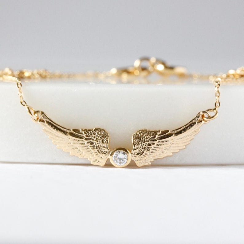 May include: A gold-colored necklace featuring a pair of detailed wings with a small, clear gemstone at the center. The wings are spread out, and the necklace has a delicate chain.
