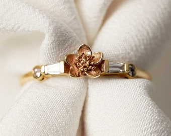 May include: A gold ring with a delicate flower design and small clear stones. The ring is on a white fabric background.