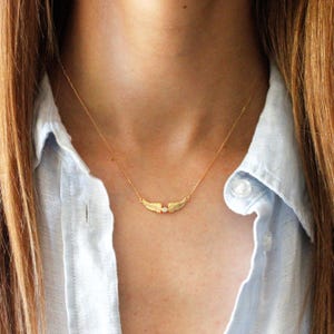 May include: A delicate gold-colored necklace featuring a pair of textured wings with a small, clear gemstone at the center. The necklace is suspended from a fine chain, creating a minimalist and elegant design. The jewelry is worn against a light blue shirt.