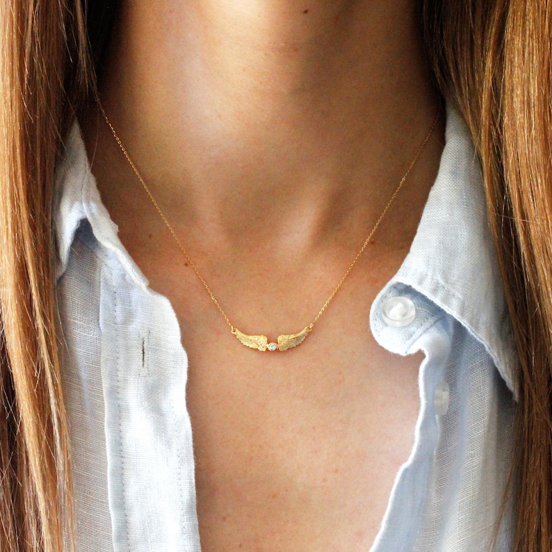 May include: A delicate gold-colored necklace featuring a pair of textured wings with a small, clear gemstone at the center. The necklace is suspended from a fine chain, creating a minimalist and elegant design. The jewelry is worn against a light blue shirt.