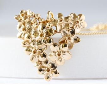 May include: A gold-colored pendant necklace featuring a cluster of detailed flower blossoms. The pendant is shaped like a heart and is suspended from a delicate gold chain. The flowers have intricate petal details and a polished finish.
