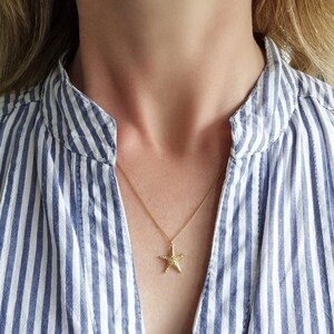 Pode incluir: Um colar de ouro com um pingente de estrela do mar texturizado. O colar &eacute; usado por uma pessoa que usa uma camisa listrada azul e branca.