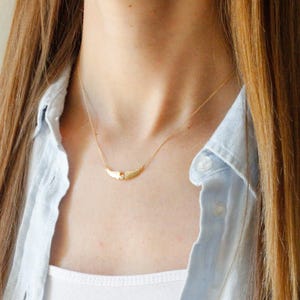May include: A delicate gold necklace featuring a pair of stylized wings with a small, clear gemstone at the center. The necklace is worn against a light blue button-down shirt and a white tank top.