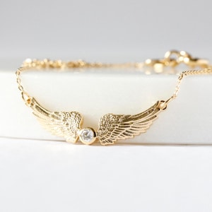 May include: A gold-colored necklace featuring a pair of detailed angel wings. A small, clear gemstone is set between the wings. The necklace chain is delicate and extends on either side of the wings, creating a graceful and elegant design.