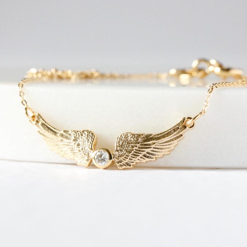 May include: A gold-colored necklace featuring a pair of detailed angel wings. A small, clear gemstone is set between the wings. The necklace chain is delicate and extends on either side of the wings, creating a graceful and elegant design.