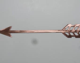 May include: A copper-colored metal arrow pointing to the left. The arrow is made of a single piece of metal and has a simple, modern design.