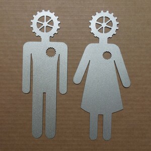 May include: Two silver metal silhouettes of people with gear-shaped heads. The silhouettes are standing on a brown surface.