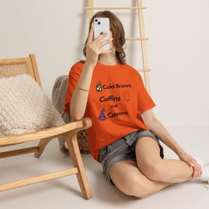 May include: An orange short-sleeved t-shirt featuring the black text Cold Brews, Coffins, and Covens. The design includes small graphic icons of a coffee cup, a coffin, and a purple witch hat with a yellow buckle, printed on the front of the shirt.