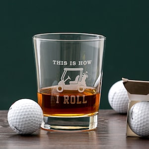 May include: A clear glass with a golf cart design and the text "This is how I roll" etched on the side. The glass is filled with amber liquid and there are golf balls in the background.