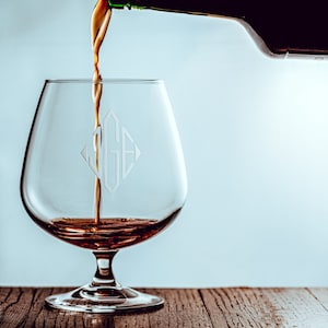May include: A clear glass brandy snifter with a monogram etched on the side is being filled with amber-colored liquid from a bottle.