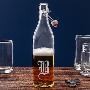 May include: A clear glass bottle with a swing top lid and a white letter "B" etched on the front. The bottle is filled with a light brown liquid and has a white head of foam on top. The bottle is sitting on a wooden surface with several bottle caps scattered around it.