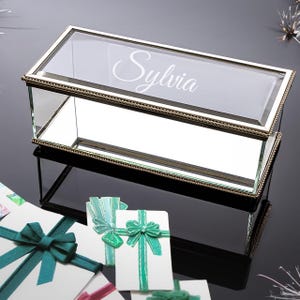 May include: A rectangular glass box with a mirrored base and a gold-colored metal frame. The lid is etched with the name "Sylvia" in a cursive font. The box is surrounded by gift boxes with teal and green ribbons.