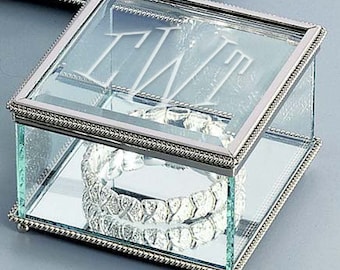 May include: A clear glass jewelry box with a silver trim and a beveled edge. The lid has an etched monogram of the letters "TW" in a script font.