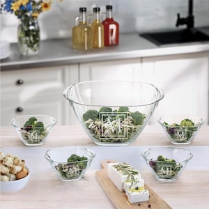 May include: A set of clear glass serving bowls, including a large bowl and several smaller bowls, each filled with salad ingredients. The bowls feature a decorative monogram. The image suggests a kitchen setting with food preparation.