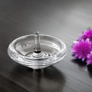 May include: A clear glass ring holder with a raised center post and a swirling design. The holder is engraved with the name "Natalie".