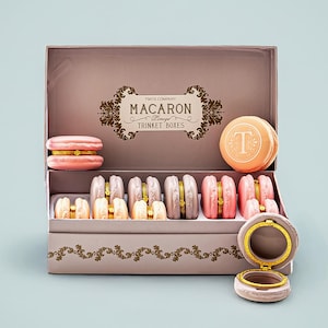 May include: A decorative box filled with macaron-shaped trinket boxes. The box is pink with gold accents and features the text "MACARON Limoges TRINKET BOXES". The trinket boxes are in various pastel colors, including pink, peach, and lavender.