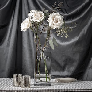 May include: A clear, rectangular glass vase with a white floral "K" monogram, holding white roses and greenery. Two mercury glass votive candle holders and a small, white plate with a gold rim are on a white textured surface.