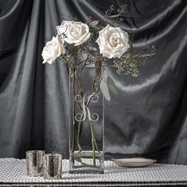 May include: A clear, rectangular glass vase with a white floral "K" monogram, holding white roses and greenery. Two mercury glass votive candle holders and a small, white plate with a gold rim are on a white textured surface.