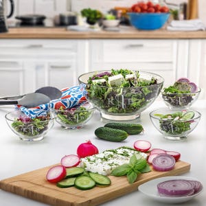 May include: A clear glass salad bowl and smaller bowls filled with salad, radishes, cucumbers, and feta cheese. The large bowl and smaller bowls have a monogram design. A wooden cutting board holds the fresh ingredients. A pair of tongs and a patterned cloth are also visible.
