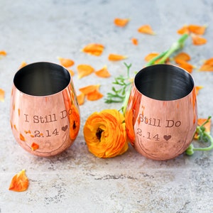 May include: Two copper-colored wine glasses with the text "I Still Do 2.14" and "Still Do 2.14" engraved on them. The glasses are sitting on a gray surface with orange flower petals scattered around them.