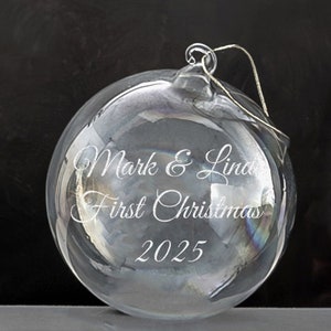 May include: A clear glass Christmas ornament with the text "Mark & Linda First Christmas 2025" etched on the surface.