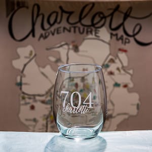 May include: A clear glass wine glass with the text "704 charlotte" etched on the side. The glass is sitting on a white surface in front of a blurred background of a map.