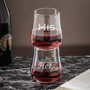 May include: Two clear glass tumblers stacked on top of each other. The top tumbler has the word "His" etched on the side, and the bottom tumbler has the word "Hers" etched on the side. Both tumblers are filled with a red liquid.
