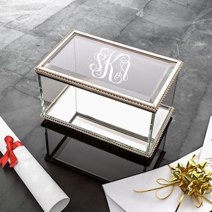 May include: A clear glass box with a mirrored bottom and a silver trim. The box has a rope-like design around the edge and is engraved with the initials "SK".