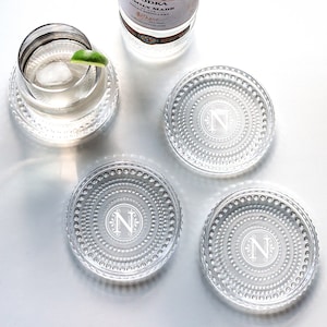 May include: Four clear glass coasters with a beaded edge and a monogrammed letter N in the center. The coasters are on a white surface.
