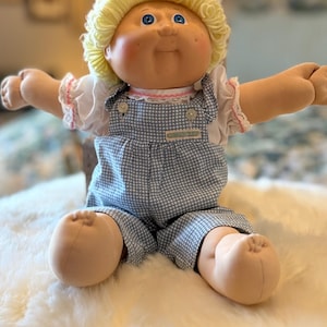 May include: A doll with light skin, blue eyes, and curly yellow yarn hair. The doll wears a white short-sleeved shirt with pink trim and blue and white checkered overalls. A small rectangular label is attached to the front of the overalls.