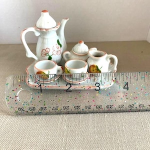 May include: Miniature porcelain tea set with a white teapot, sugar bowl, creamer, two teacups, and a serving tray. The set is decorated with pink floral and fruit designs. The tray measures approximately 4 inches in length.