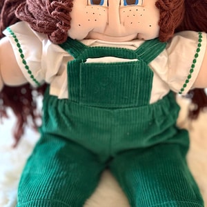 May include: A soft-sculpture doll with brown yarn hair tied in pigtails with pink beads, blue eyes, and freckles. The doll wears a white short-sleeved shirt with green trim and dark green corduroy overalls with a front pocket and wide legs.