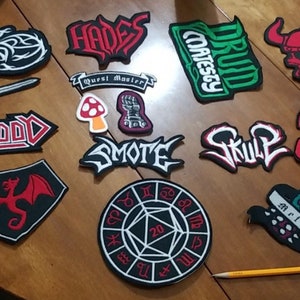 May include: A collection of 12 embroidered patches featuring various designs, including a skull, a dragon, a mushroom, a sword, and a 20-sided die. The patches are all different colors and have a variety of text, including "Hades", "Smote", "War Axe", and "Quest Master".