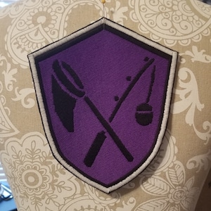 May include: Purple embroidered patch with a white border in the shape of a shield. The patch features two crossed tools, a fishing rod and a net, in black.