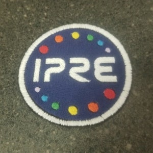 Puede incluir: Un parche circular azul con costuras blancas alrededor del borde. El centro del parche tiene el texto "IP2E" en letras blancas. El parche está decorado con un círculo de puntos de colores alrededor del borde.