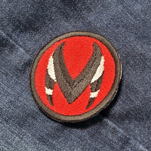 Puede incluir: Un parche bordado rojo, negro y blanco con una letra estilizada 'M' en el centro. El parche es circular y tiene un borde negro.